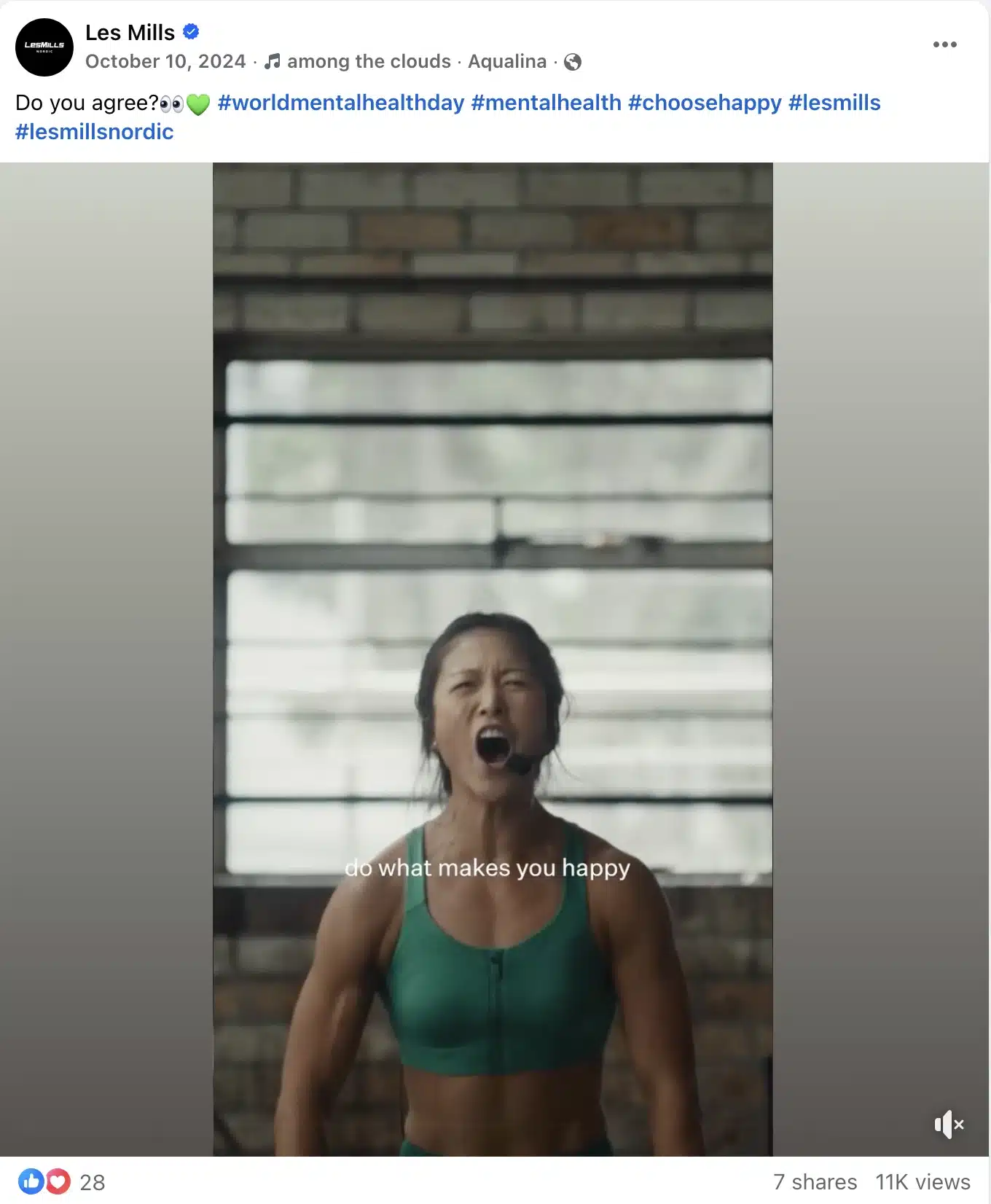 Les Mills fitness instructor wearing a green sports bra and headset microphone, mid-workout in a studio with brick walls and large windows, with on-screen text reading “do what makes you happy,” promoting mental health and movement.