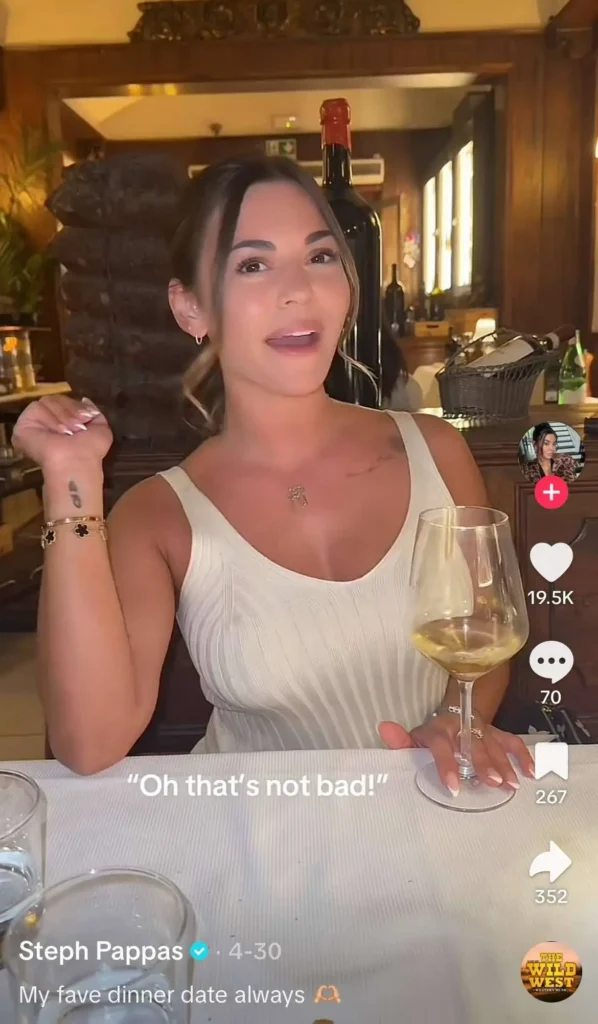 A woman sitting at a dining table in a restaurant, holding a glass of white wine. Text overlay reads: "Oh that's not bad!"