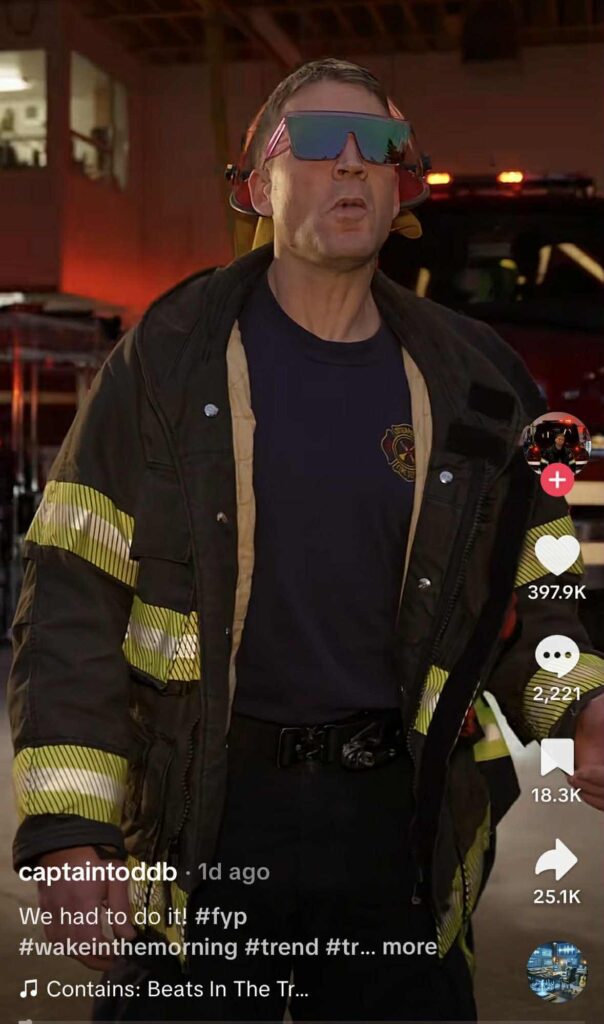 A firefighter wearing reflective gear and large mirrored sunglasses stands in front of a fire truck with flashing lights, participating in a TikTok trend.
