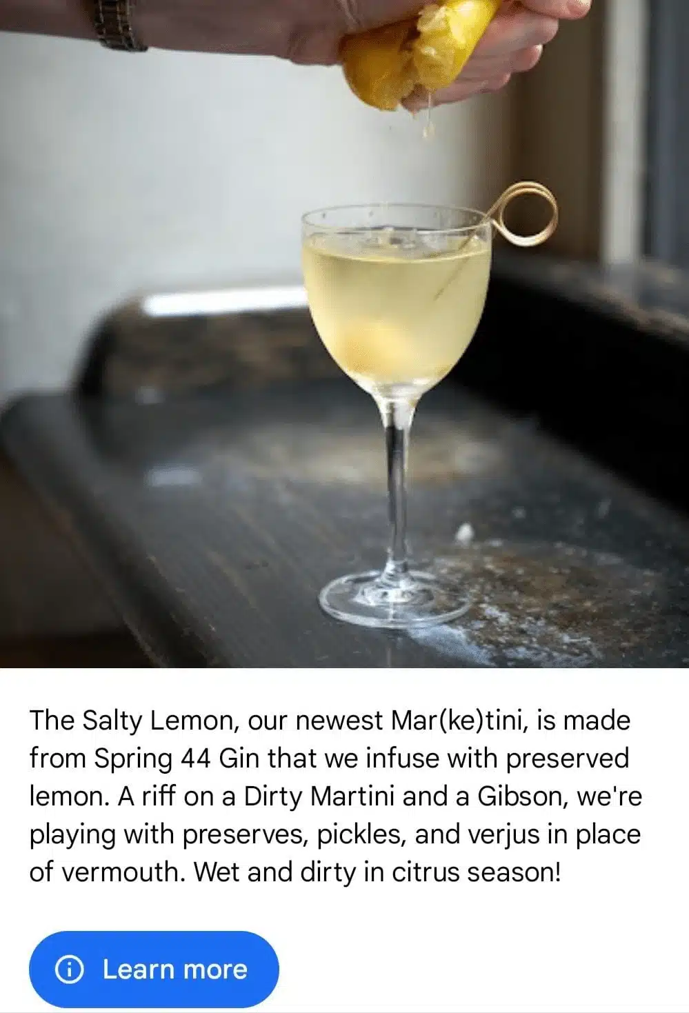 A hand squeezes a lemon over a chilled martini glass containing The Salty Lemon cocktail, featured in a Google Business Profile post promoting a gin-based drink infused with preserved lemon and garnished with a cocktail pick.