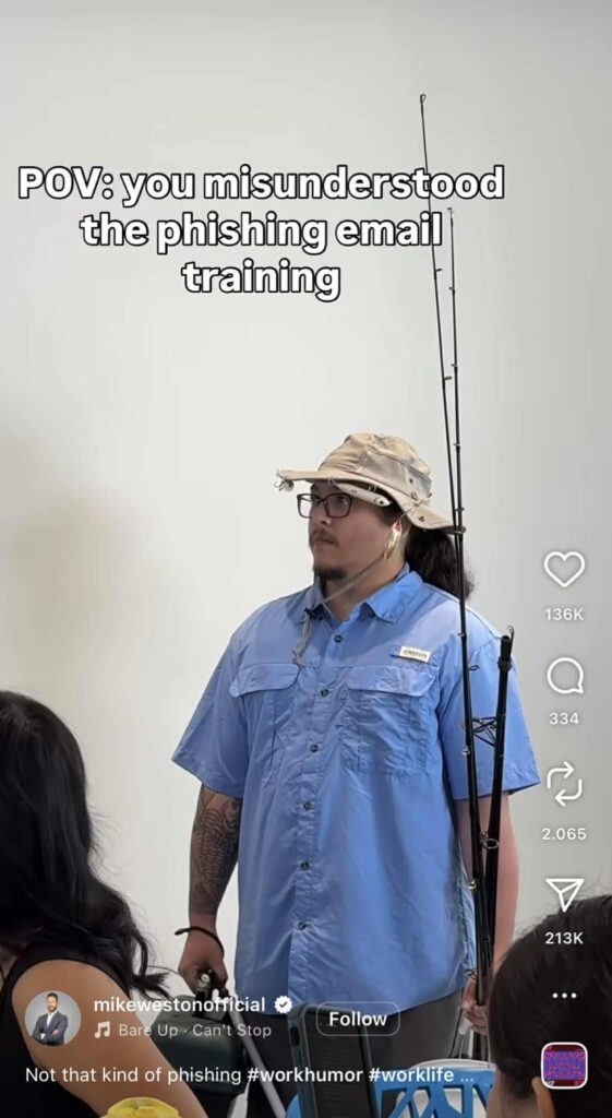 A person is standing indoors wearing a light blue fishing shirt and a beige bucket hat, holding a tall fishing rod. The background shows a white wall and part of a seated audience. Text at the top of the image reads: “POV: you misunderstood the phishing email training.”