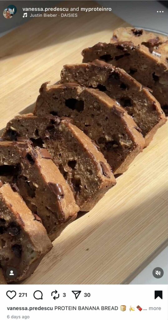 Close-up of sliced protein banana bread with melted chocolate chunks on a wooden board, shared in an Instagram Reel.