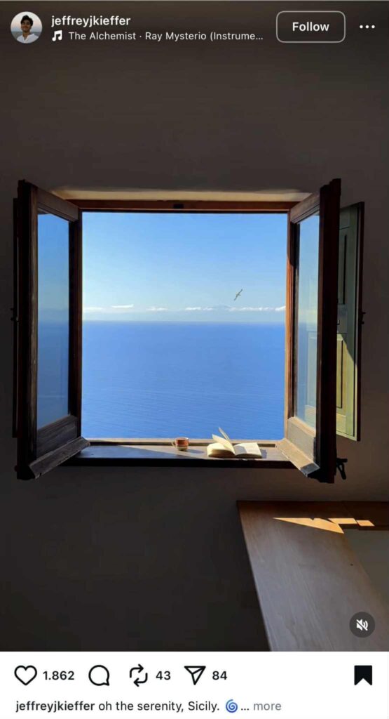 Instagram Reel showing an open wooden window overlooking the calm blue sea in Sicily. A cup of coffee and an open book sit on the windowsill, with a bird flying in the distance under a clear sky.