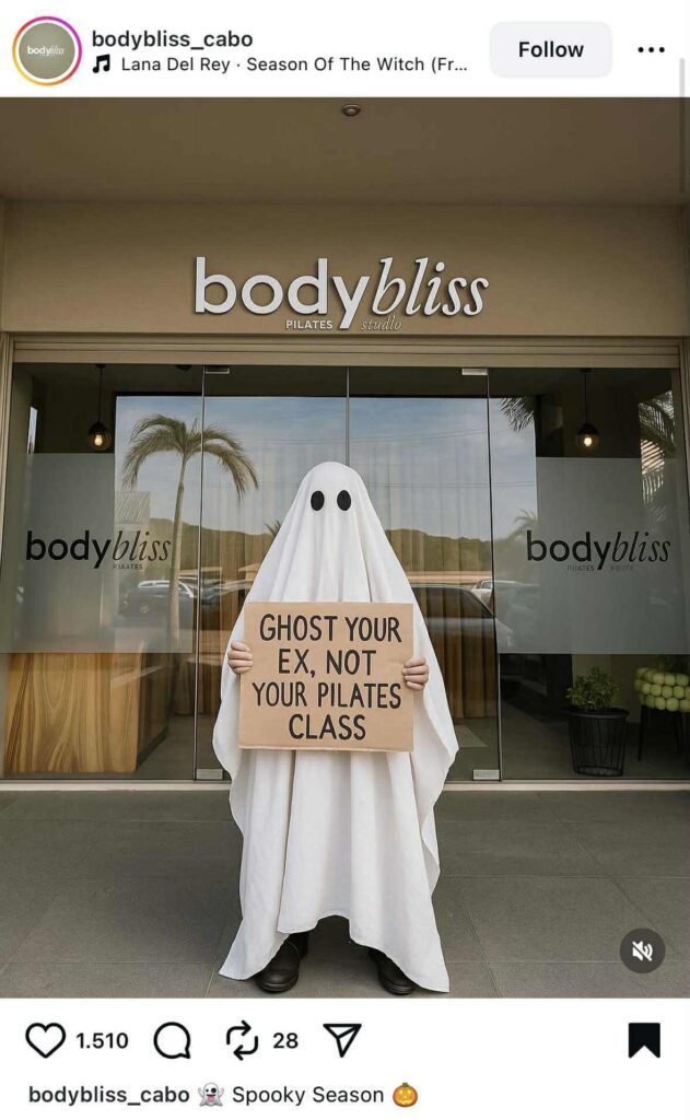 A person dressed as a ghost stands in front of a storefront with the sign “bodybliss PILATES studio.” The ghost is holding a cardboard sign that reads, “GHOST YOUR EX, NOT YOUR PILATES CLASS.” 