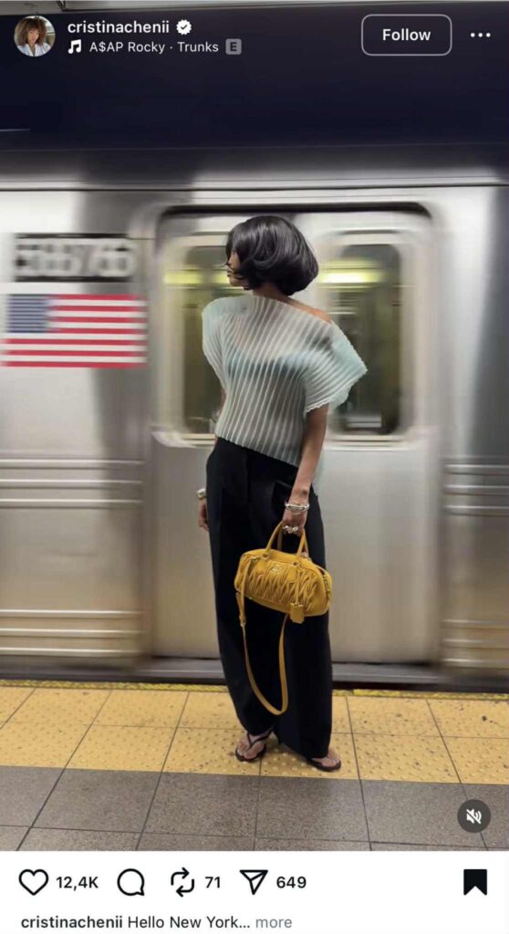 Instagram Reel of a woman standing on a subway platform in New York. She wears a pleated off-shoulder top, black pants, and sandals, holding a mustard-yellow handbag. A subway train blurs past in the background with the American flag visible.