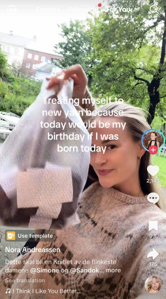 A woman smiles while holding up a shopping bag filled with yarn outdoors. The on-screen text reads, “Treating myself to new yarn because today would be my birthday if I was born today.”
