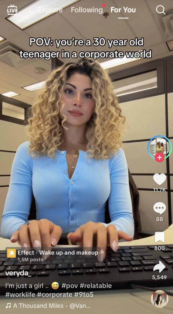 A woman with curly blonde hair sits at her desk in an office, typing on a keyboard and looking at the camera with a confident expression. The TikTok caption reads, “POV: you’re a 30 year old teenager in a corporate world.”