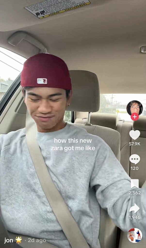 TikTok of a person sitting in a car seat wearing a red cap and gray sweatshirt, smiling and dancing with eyes closed. The on-screen text reads, “how this new zara got me like.”