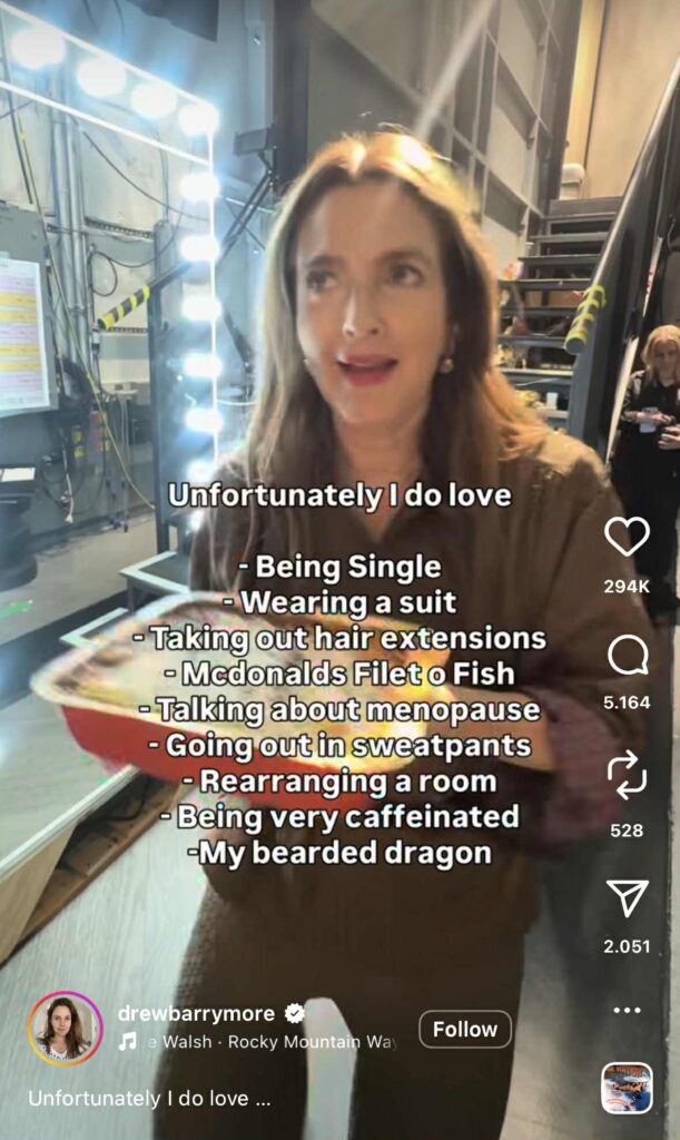 Instagram Reel of Drew Barrymore walking backstage holding food while text on screen lists things she loves. The caption starts with “Unfortunately I do love” followed by a humorous list including being single, wearing a suit, McDonald’s Filet-O-Fish, talking about menopause, going out in sweatpants, rearranging a room, being very caffeinated, and her bearded dragon.