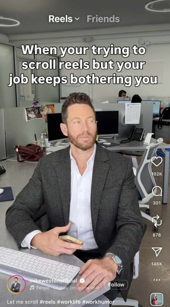 A man in a suit sits at his desk in an office holding his phone, looking slightly annoyed. The caption on the screen reads, “When you’re trying to scroll reels but your job keeps bothering you.” Computer monitors and office supplies are visible in the background.