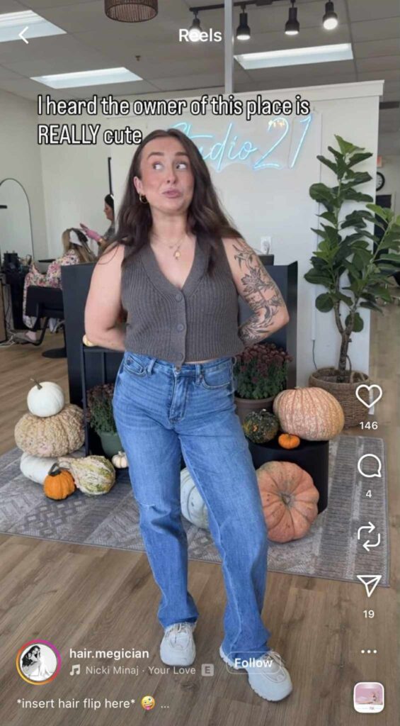 A woman standing in a hair salon smirks and looks off to the side with an amused expression. She wears a brown knit vest, blue jeans, and white sneakers, with tattoos on her arm. Behind her are fall decorations, including pumpkins and plants, and a neon “Studio 21” sign. The on-screen text reads, “I heard the owner of this place is REALLY cute.”
