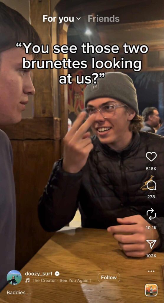 Two young men sit at a wooden table in a restaurant. One wears a beanie and glasses, smiling and pointing with two fingers toward his eyes while saying, “You see those two brunettes looking at us?” as shown in the caption.