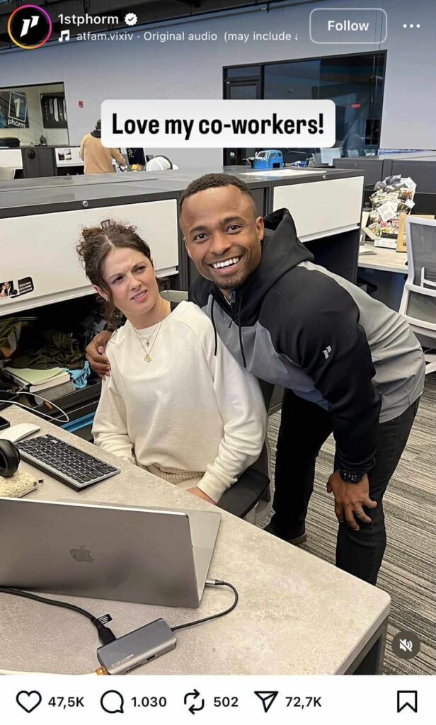 A man smiles and leans in for a cheerful photo with his coworker, who looks confused and slightly annoyed while sitting at her desk with a laptop. The caption above reads, “Love my co-workers!”