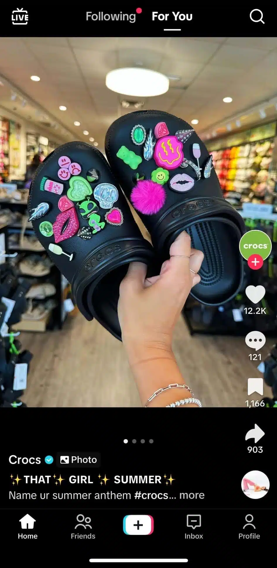 TikTok photo post by Crocs featuring a hand holding a pair of black Crocs decorated with colorful charms including smiley faces, hearts, aliens, and lips. The caption reads “✨THAT✨ GIRL ✨ SUMMER✨” with hashtags. TikTok interface icons and engagement metrics are visible on the right.