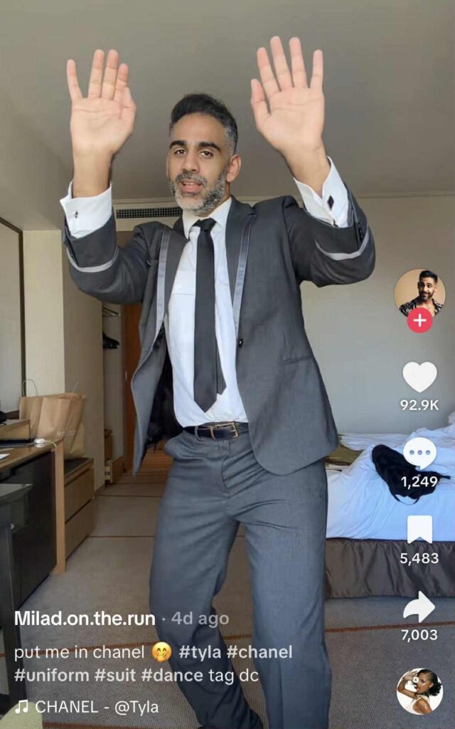 A man wearing a gray suit and black tie dances in a bedroom, raising his hands mid-move. The TikTok caption reads “put me in chanel 😚 #tyla #chanel #uniform #suit #dance tag dc,” with the song “CHANEL” by Tyla playing in the background.