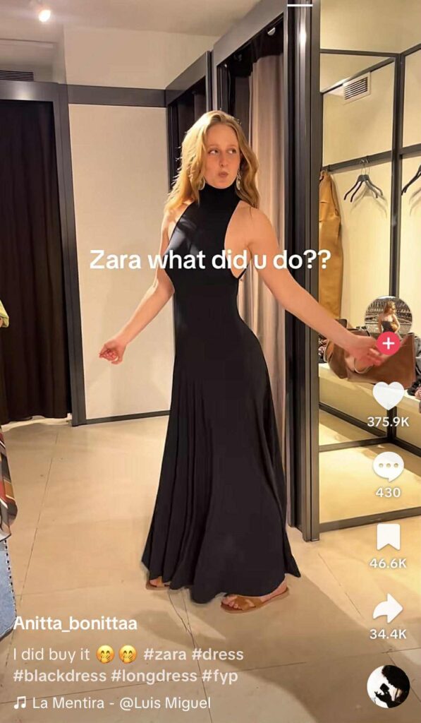 A woman poses in a fitting room wearing a long black halter dress, looking over her shoulder with a playful expression. The TikTok text overlay reads “Zara what did u do??” and the caption says “I did buy it 🥺🥺 #zara #dress #blackdress #longdress #fyp,” with the song “La Mentira” by Luis Miguel playing.