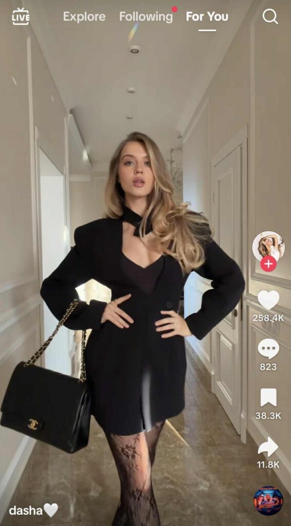 A woman poses confidently in a hallway wearing a black blazer dress, lace tights, and a Chanel handbag with a gold chain strap.