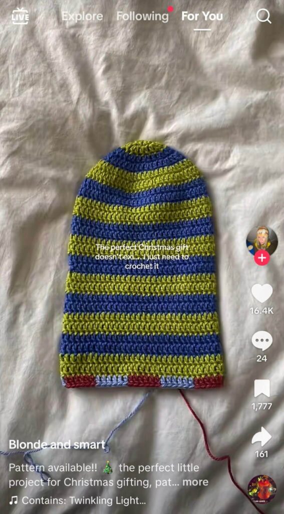 A colorful striped crocheted hat-in-progress laying on a bed with the text “The perfect Christmas gift doesn't exi... I just need to crochet it.”