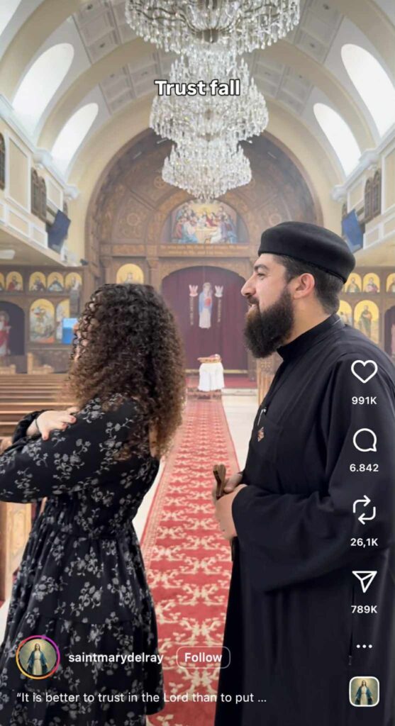 Inside a large, ornate church, a woman with curly hair stands with her arms crossed, leaning backward as if doing a trust fall. Beside her, a priest in traditional black clerical clothing watches calmly. A grand chandelier hangs above them, and religious icons and paintings decorate the background. The on-screen caption reads “Trust fall.”
