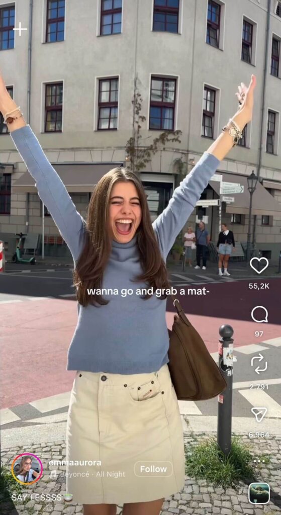 A young woman stands in a city street with her arms raised, smiling excitedly. On-screen text reads, “wanna go and grab a mat-”.
