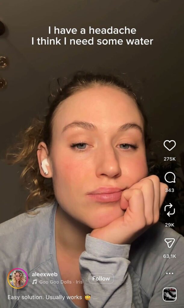 A young woman rests her chin on her hand, looking tired at the camera. On-screen text reads, “I have a headache I think I need some water.”