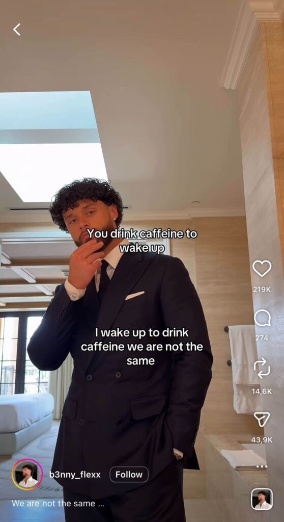 A man in a suit stands in a bathroom, touching his chin thoughtfully. Text reads, “You drink caffeine to wake up. I wake up to drink caffeine. We are not the same.”