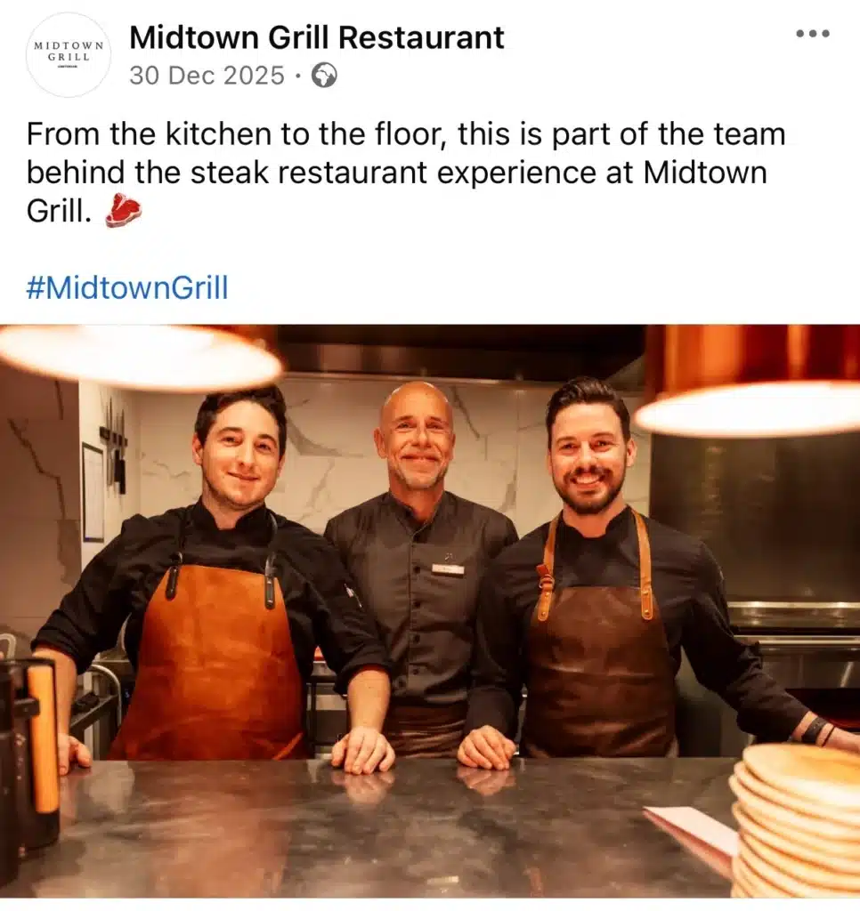 Three restaurant team members standing behind a counter in a professional kitchen, smiling at the camera as part of a behind-the-scenes team post for Midtown Grill.