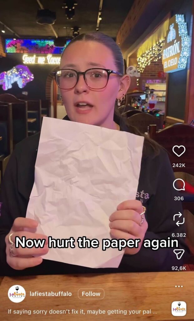 Instagram Reel of a woman holding up a crumpled sheet of paper in a restaurant setting. On-screen text reads, “Now hurt the paper again.” Instagram likes, comments, and share icons appear along the right side.
