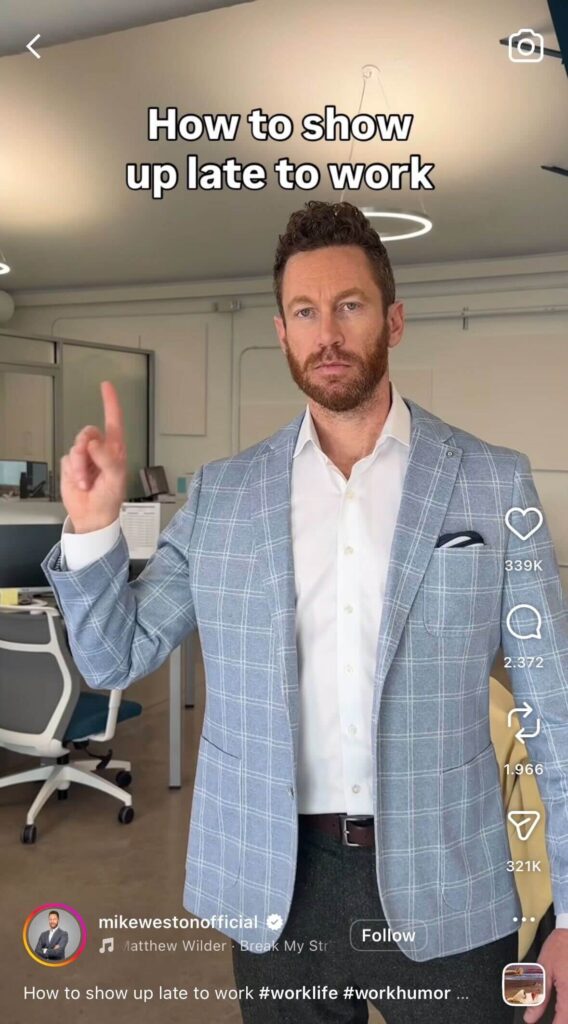 A man in a light blue blazer stands in an office pointing upward with one finger. On-screen text reads, “How to show up late to work.”