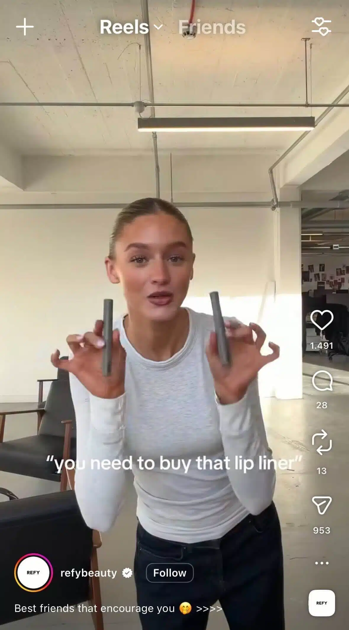 A woman in a bright studio space holds up two lip liner products toward the camera, smiling. On-screen text reads, “you need to buy that lip liner,” with a beauty brand account shown.