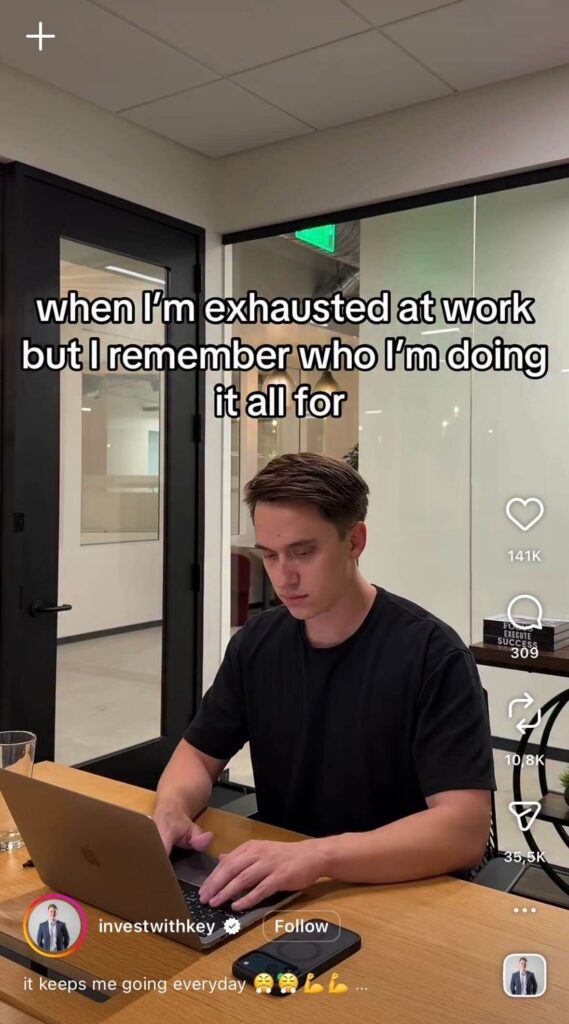A tired-looking man sits at a desk typing on a laptop in a modern office. On-screen text reads, “when I’m exhausted at work but I remember who I’m doing it all for.”