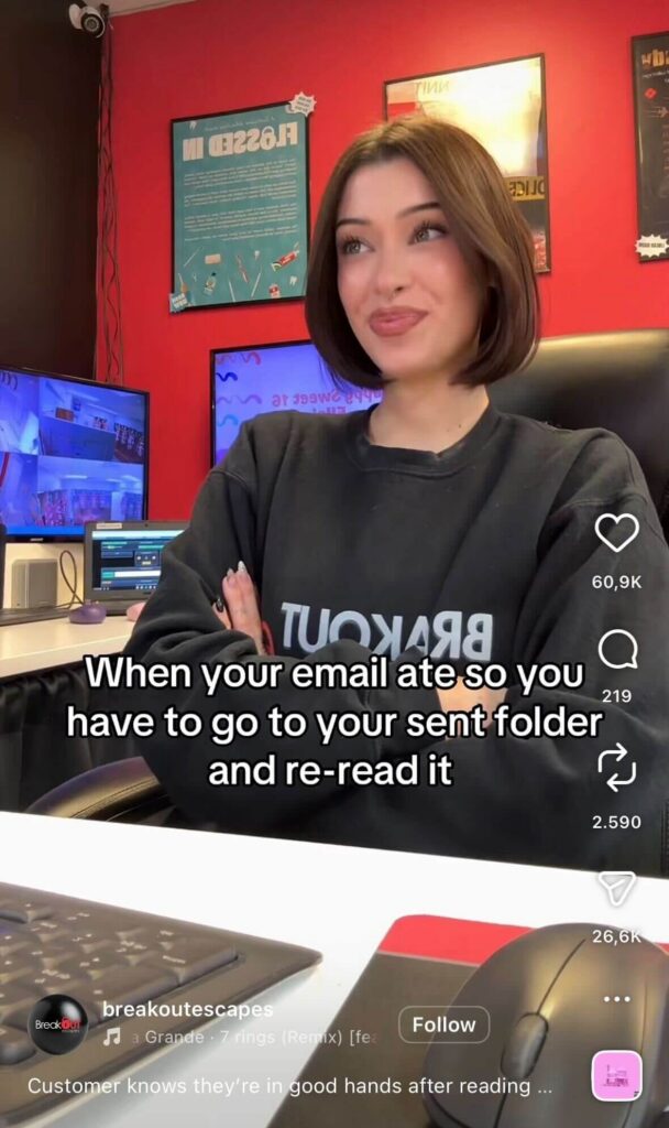 Young woman with short brown hair sitting at a desk in an office with red walls and multiple monitors, smiling with arms crossed; text reads “When your email ate so you have to go to your sent folder and re-read it.”