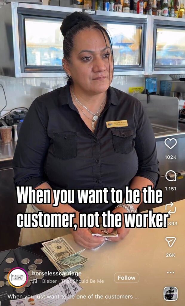 Woman in a black uniform standing behind a restaurant counter handling cash and coins, looking slightly tired; overlaid text reads “When you want to be the customer, not the worker.”