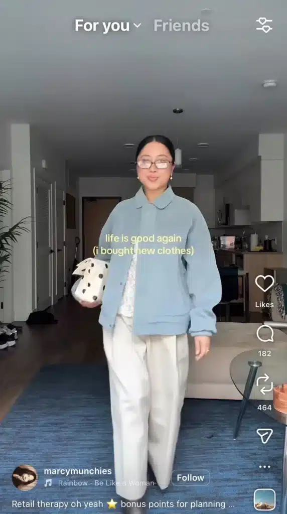 Woman wearing glasses and a light blue jacket walking through a modern apartment while holding a white polka-dot bag; overlaid text reads “life is good again (i bought new clothes).”