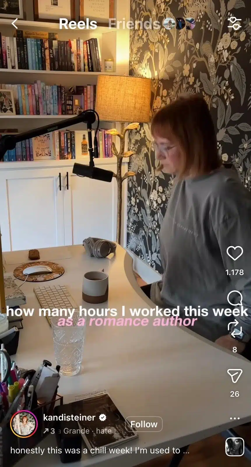 A woman sits at a curved white desk in a home office. The workspace includes a microphone on a boom arm, headphones, a keyboard, books, pens, a glass of water, and a coffee mug. Behind her are bookshelves filled with books, a floor lamp, and floral-patterned wallpaper. White and pink overlay text reads, “how many hours I worked this week as a romance author.” Instagram Reels interface elements are visible.