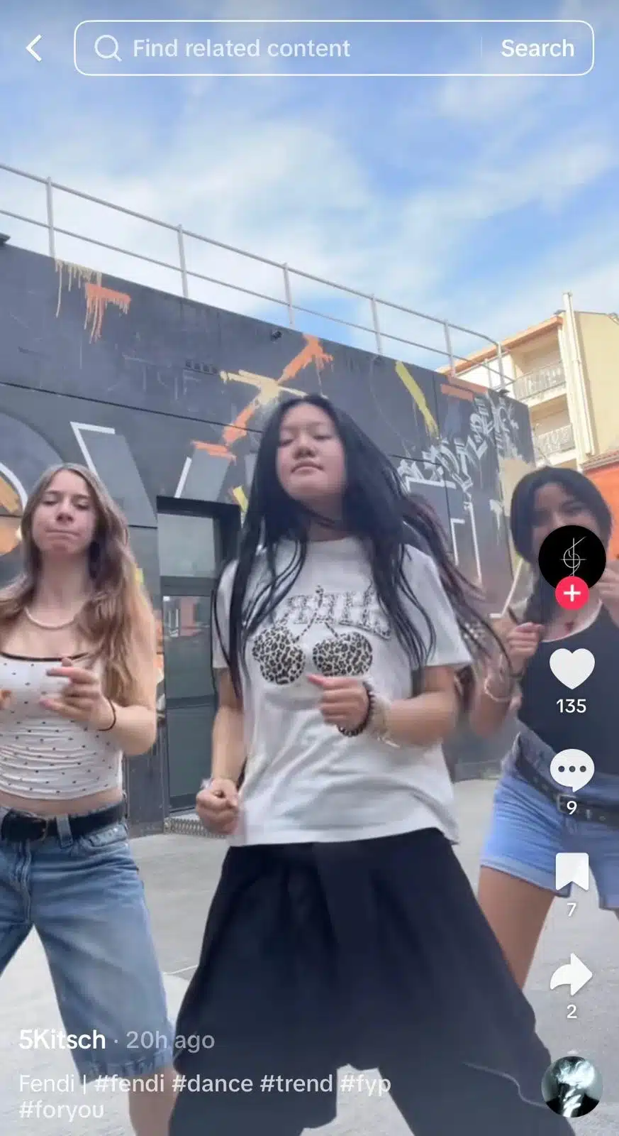 Three people dancing outdoors in front of a graffiti-covered wall. The person in the center wears a white graphic T-shirt and black skirt, while two others dance on either side. TikTok interface visible.