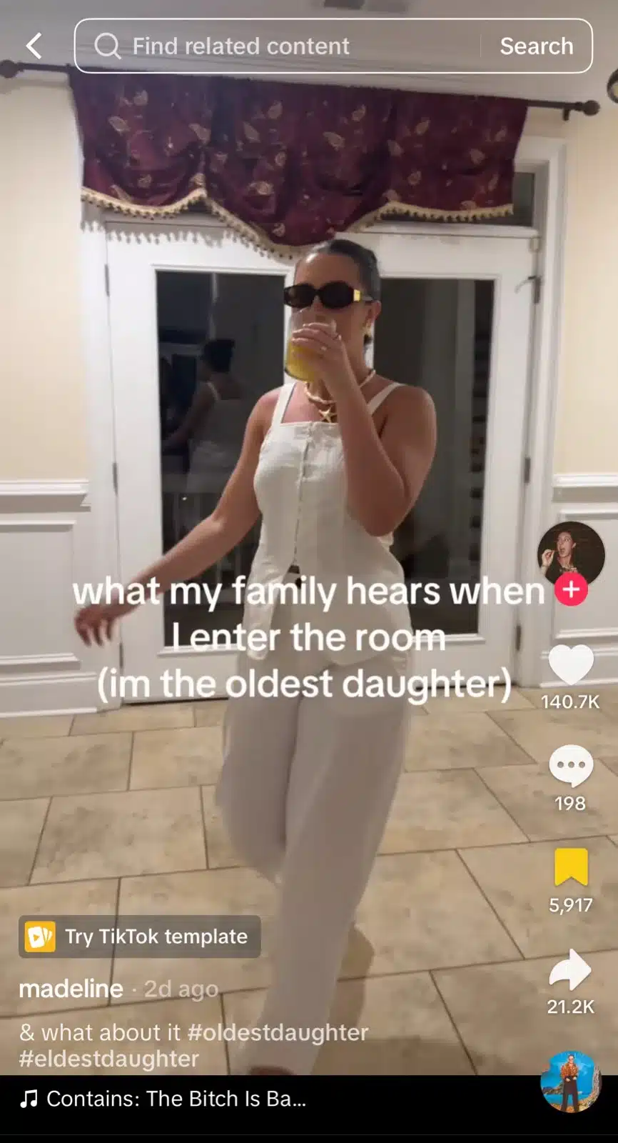 A woman dressed in a white sleeveless jumpsuit, dark sunglasses, and gold jewelry walks through a tiled room while holding a drink. Large white text over the video reads, “what my family hears when I enter the room (im the oldest daughter).”