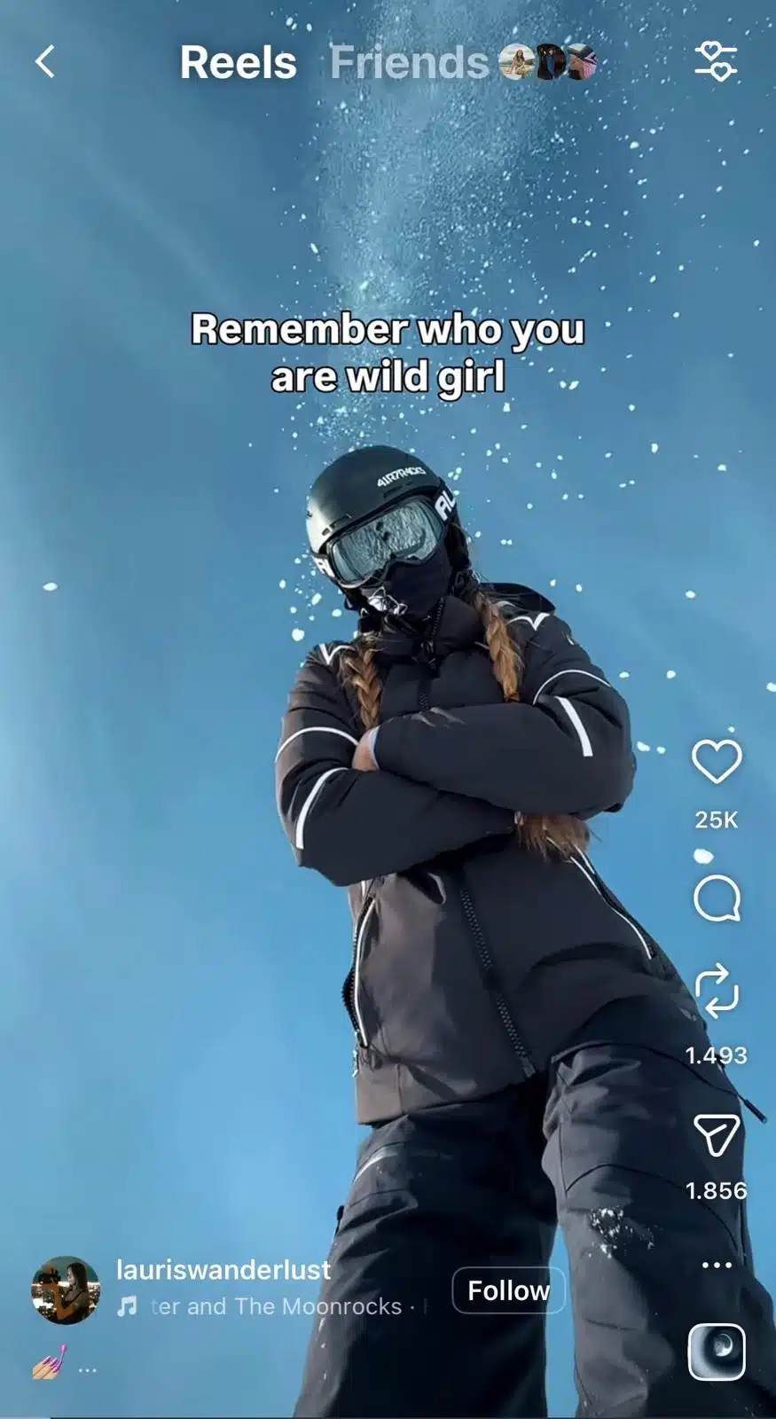 A person dressed in black ski gear stands with arms crossed while looking down at the camera. They are wearing a helmet, ski goggles, and a face covering, with braided hair visible beneath the helmet. Snow sprays into the air above them against a bright blue sky. White overlay text reads, “Remember who you are wild girl.” Instagram Reels interface elements are visible on the screen.