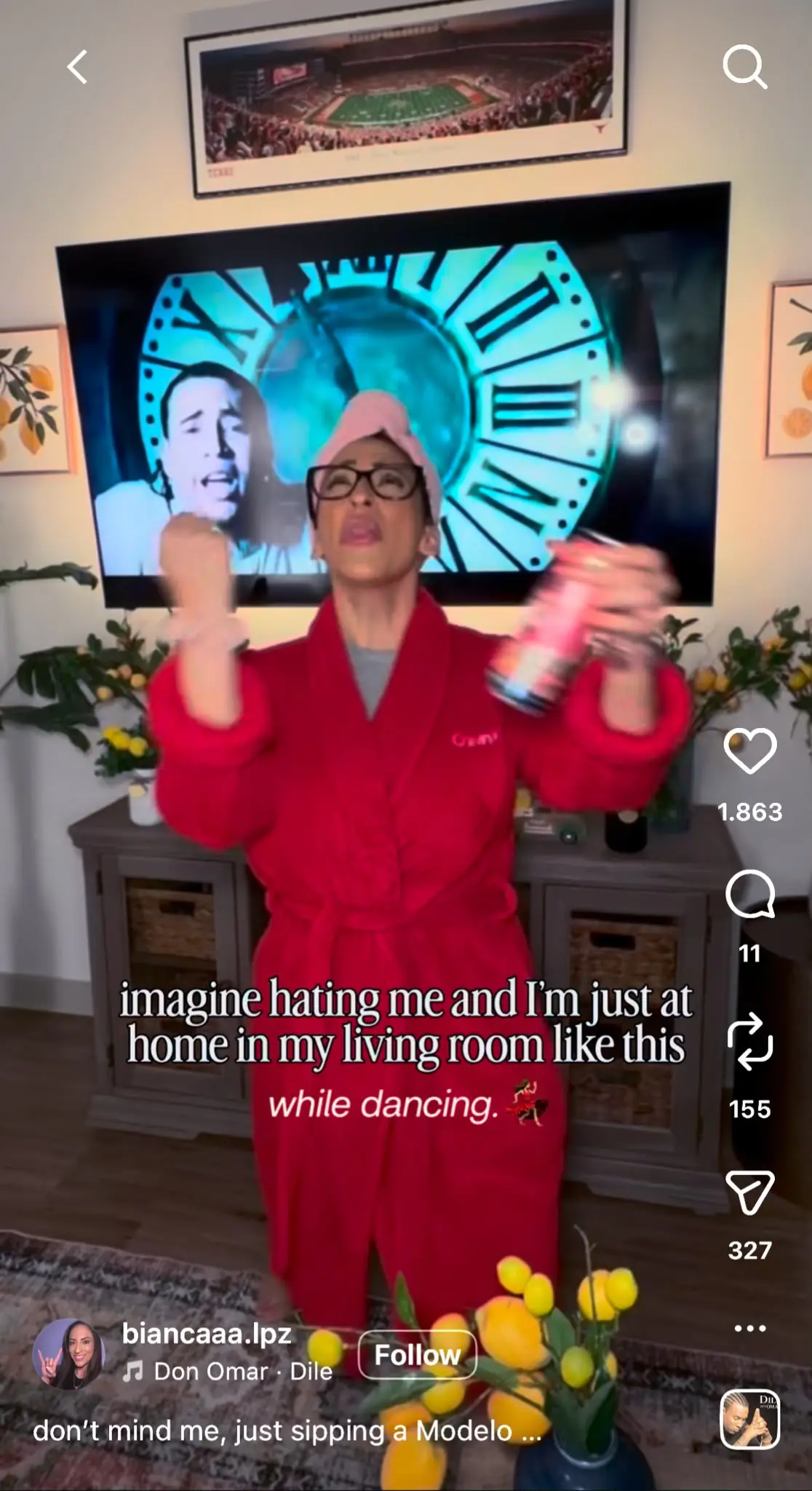 A person wearing a bright red robe, pink cap, and glasses dances enthusiastically in a living room while holding a drink can. A television behind them displays a large clock face. Overlay text reads, “imagine hating me and I’m just at home in my living room like this while dancing.”