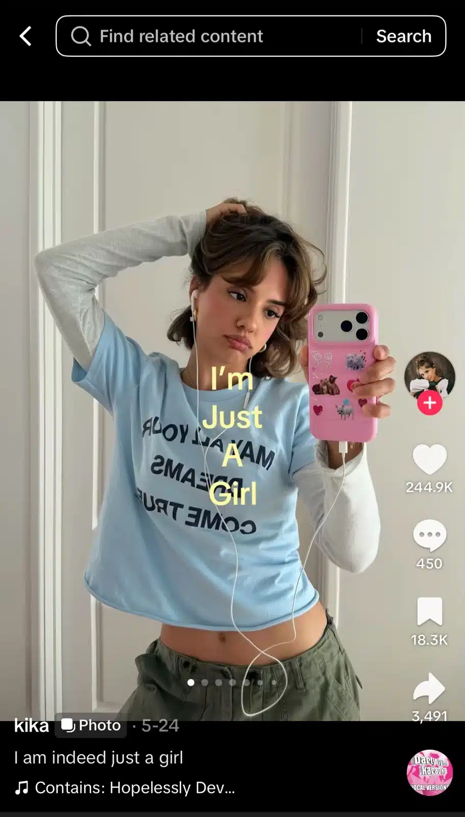 A young woman takes a mirror selfie while wearing a light blue graphic T-shirt layered over a long-sleeve white shirt and olive-green pants. She holds a pink phone case decorated with stickers and wears wired earphones. Yellow text over the image reads, “I’m Just A Girl.”