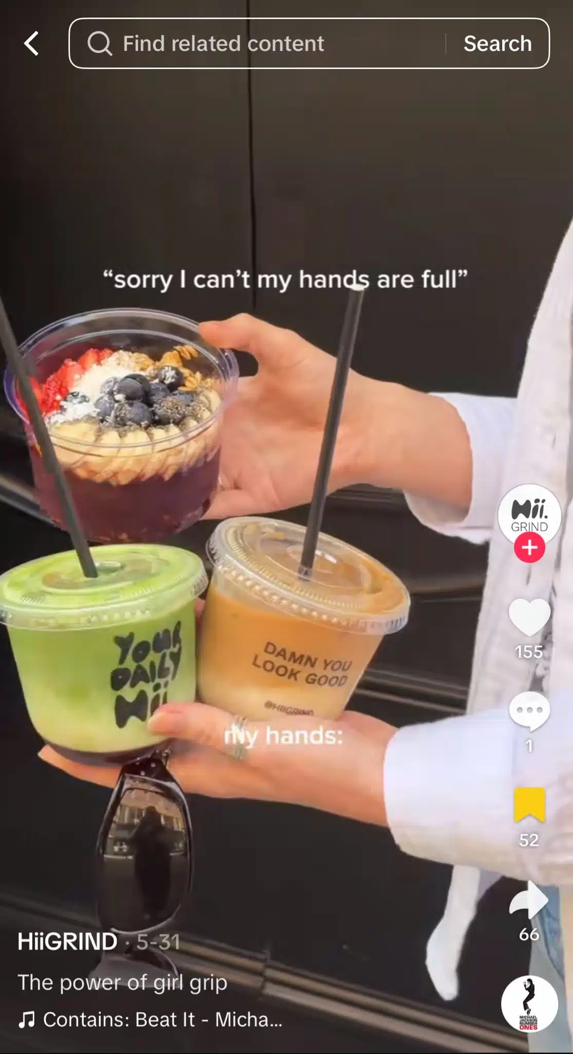 A person balances an acai bowl, two iced drinks, and a pair of sunglasses in their hands in front of a dark storefront. Overlay text reads, “sorry I can’t my hands are full” and “my hands:” while showing the items being carried.