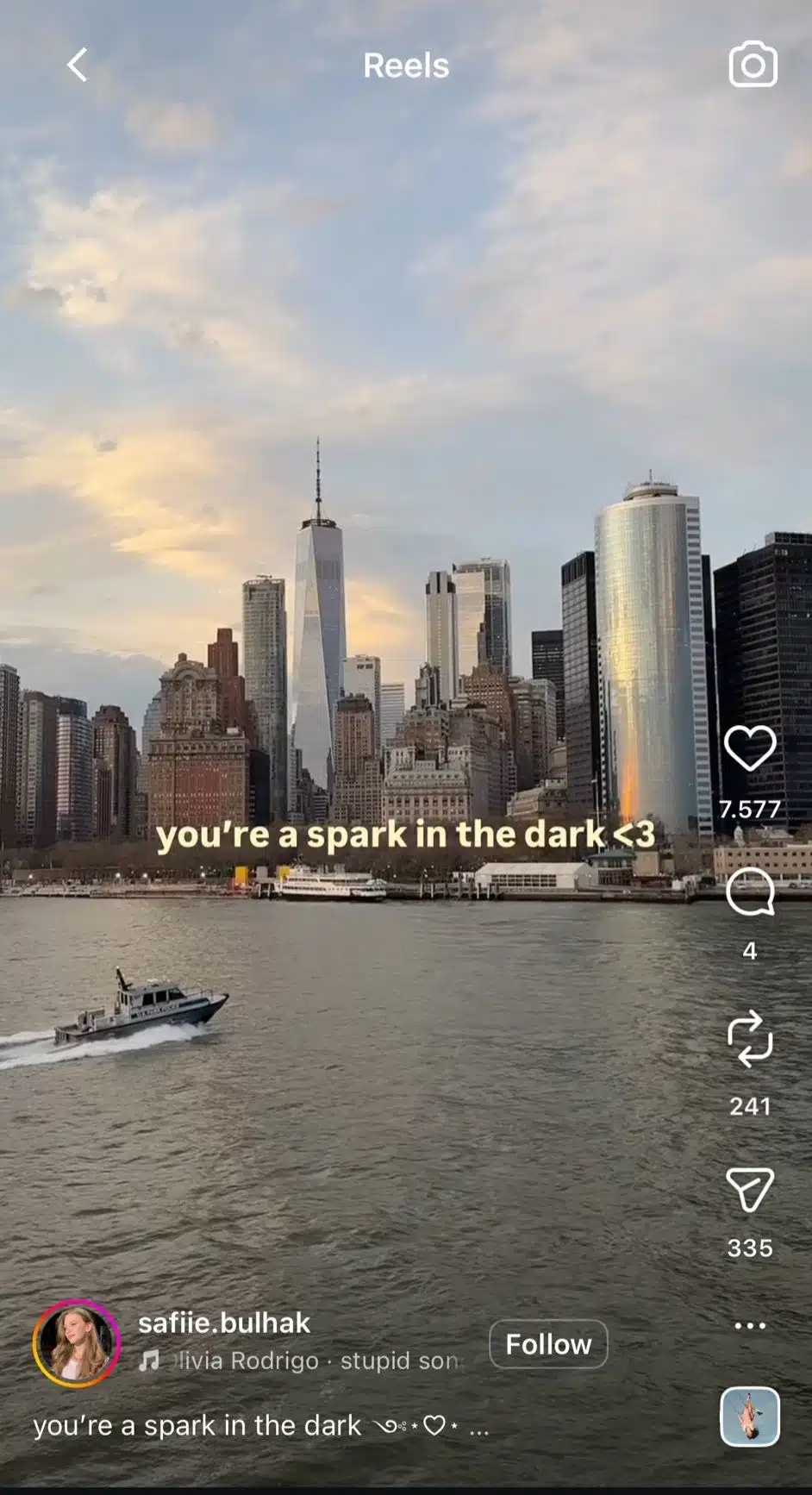 View of the Lower Manhattan skyline and One World Trade Center across a river at sunset, with a small boat traveling on the water in the foreground. On-screen text reads, “you’re a spark in the dark <3”.