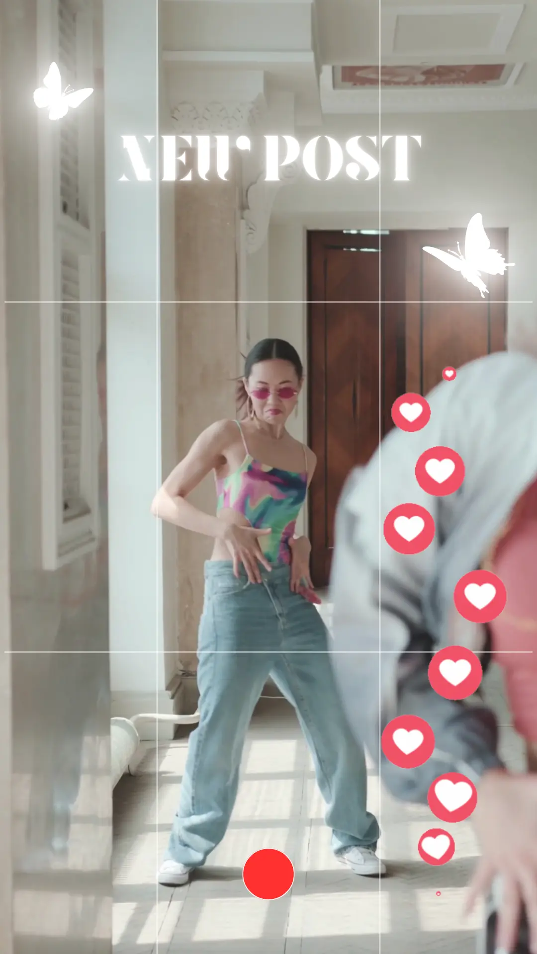 Una maqueta vertical de una historia de redes sociales en la que se ve a una mujer bailando en un pasillo luminoso mientras la graban. En la parte superior, un texto en blanco dice «NUEVA PUBLICACIÓN», con gráficos decorativos de mariposas e iconos de reacciones en forma de corazones flotantes a lo largo del lado derecho. Las líneas de cuadrícula y un botón de grabación superpuesto imitan la interfaz de la cámara de una red social.