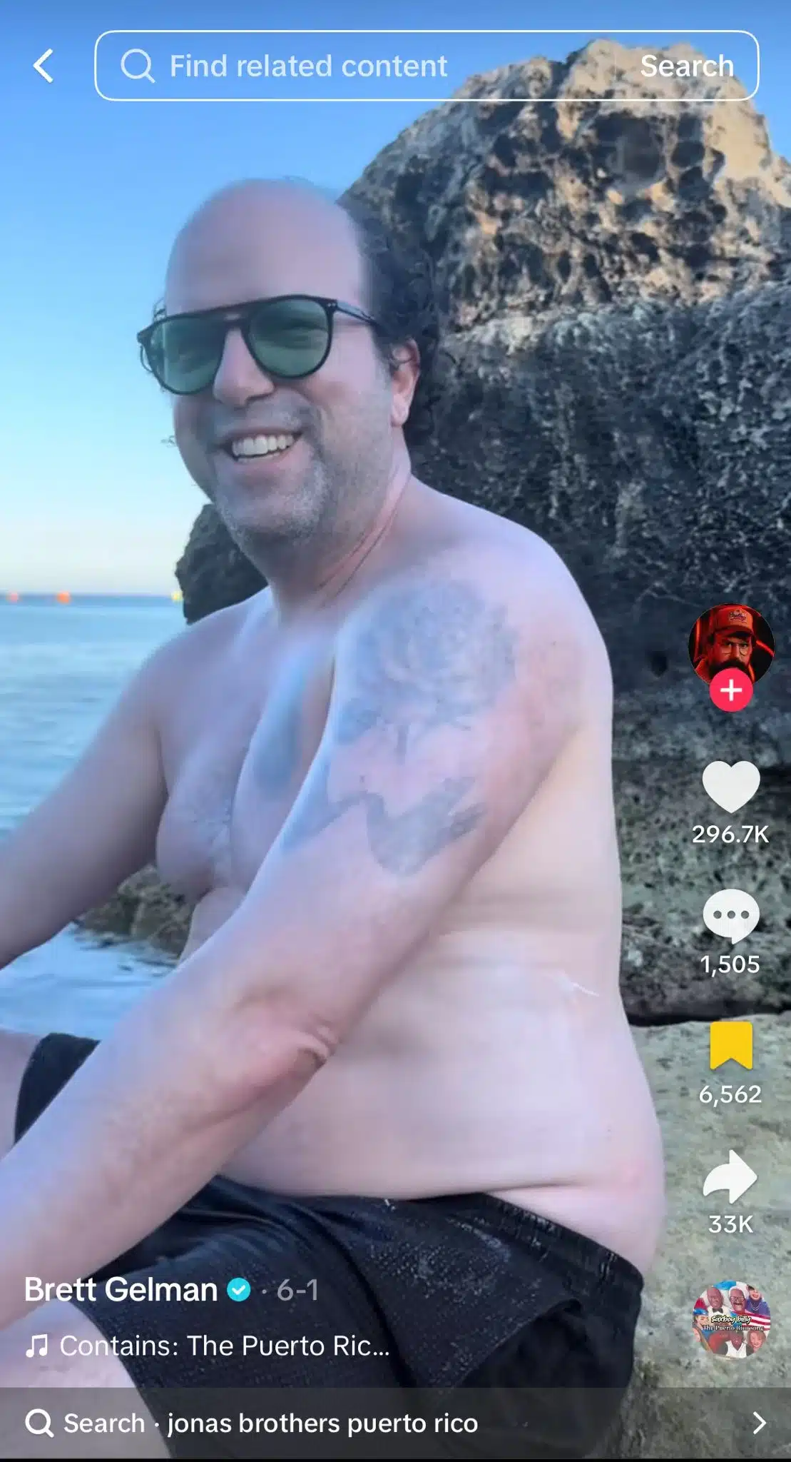 Person wearing sunglasses sitting shirtless on a rocky beach beside the water. Large rock formation behind them and ocean visible in the background. TikTok interface is visible.