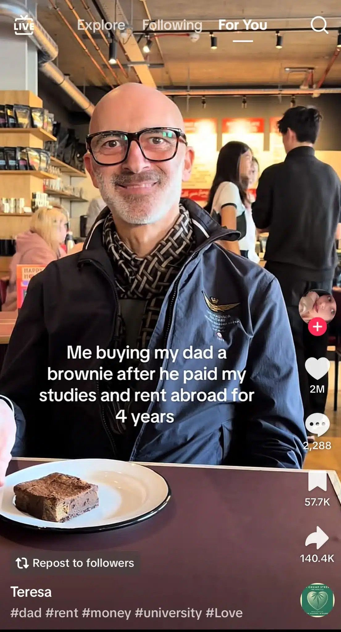 A man wearing glasses, a dark jacket, and a patterned scarf sits at a café table with a brownie on a white plate in front of him. The background shows a busy coffee shop interior with customers and menu boards. White overlay text reads, “Me buying my dad a brownie after he paid my studies and rent abroad for 4 years.” TikTok interface elements are visible.