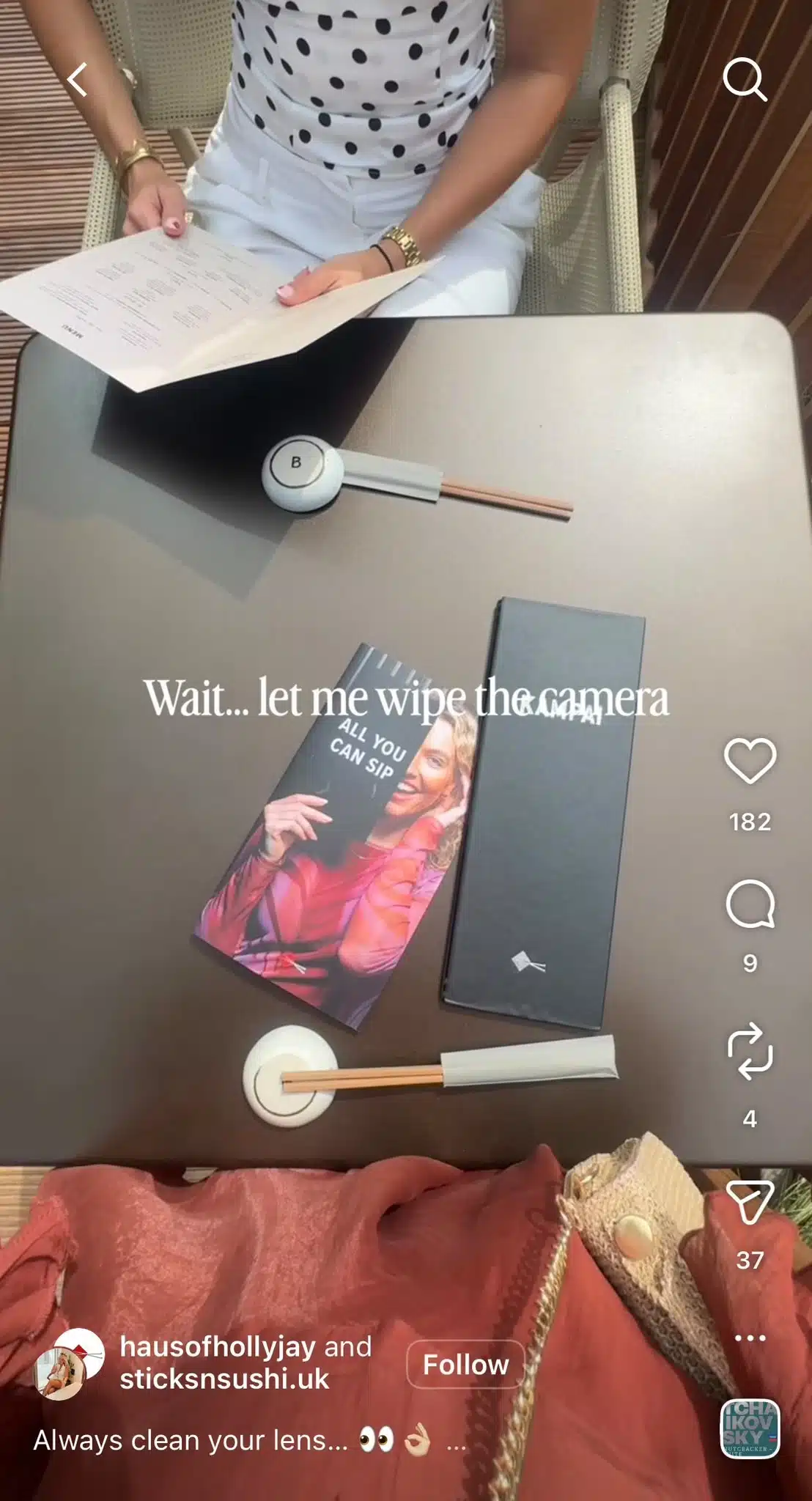 A restaurant table is viewed from above, with menus, chopsticks, and promotional materials laid out on the surface. A woman wearing a white outfit with black polka dots sits across the table holding a menu. White overlay text reads, “Wait... let me wipe the camera.”