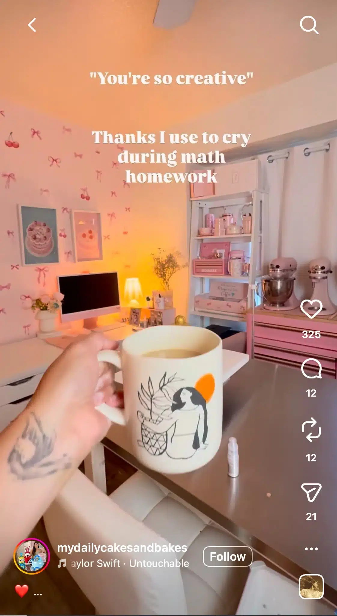 A hand holds a ceramic mug in front of a cozy pink-themed home office featuring a desk, computer, shelving, decorative wall art, and baking equipment. Warm lighting fills the room. White overlay text reads, “'You're so creative' Thanks I use to cry during math homework.”
