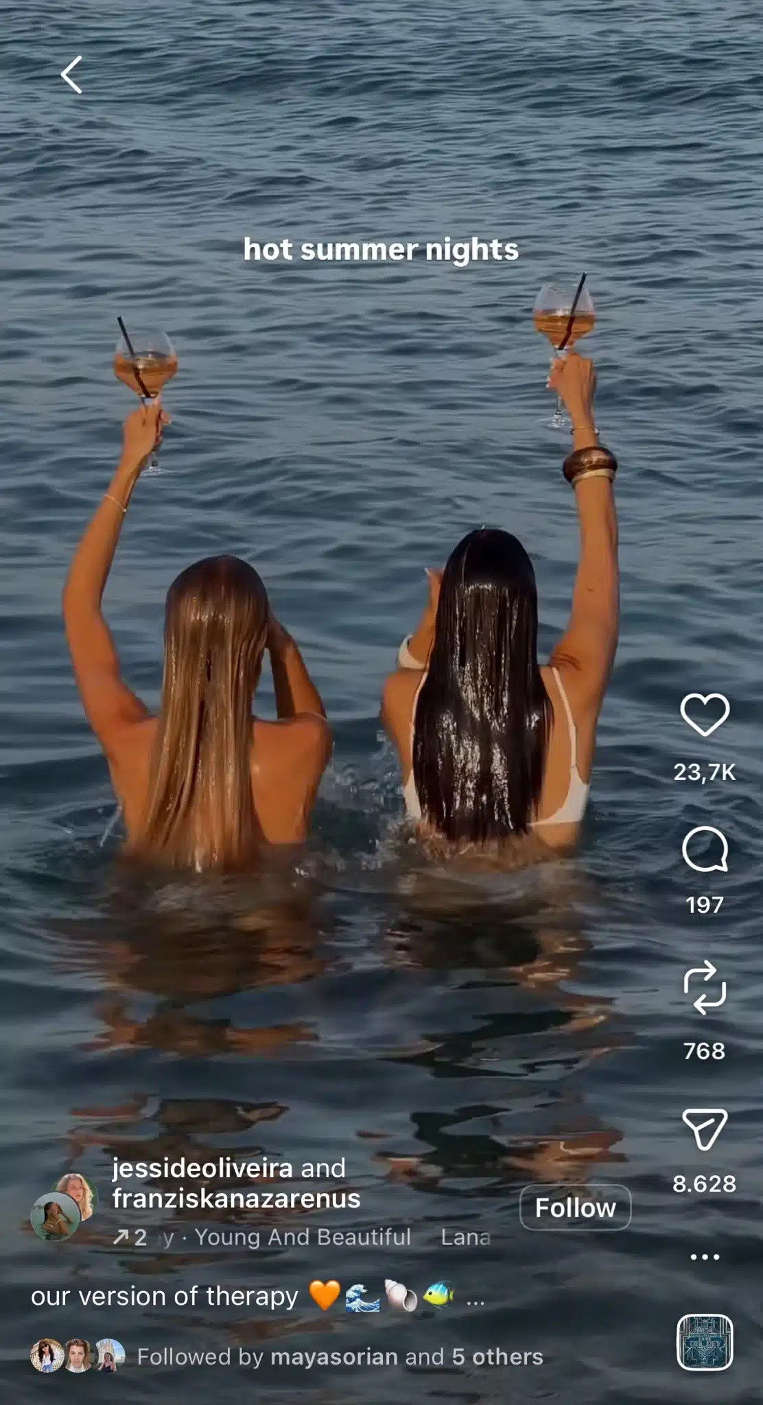 Instagram Reel showing two people standing waist-deep in the sea at sunset, each raising a wine glass while the on-screen text reads "hot summer nights." The Reel uses the "Young and Beautiful" audio trend to capture a carefree summer evening and nostalgic vacation aesthetic.