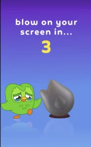 Duolingo mascot standing beside a gray flame-shaped object on a purple-to-blue gradient background, with the text “blow on your screen in... 3”.