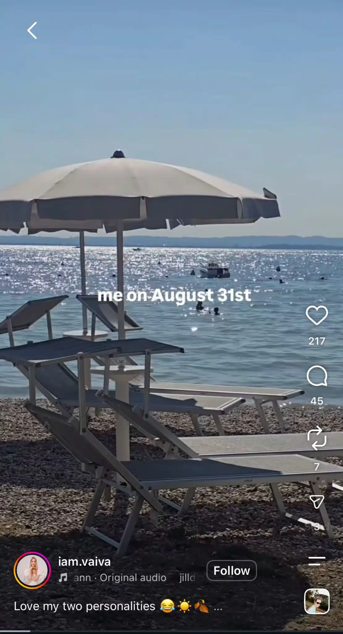 An Instagram-style Reel showing a sunny pebble beach with lounge chairs, umbrellas, sparkling blue water, swimmers, and a small boat in the distance. White text reads “me on August 31st.” The account name and Reel engagement controls appear along the bottom and right side.